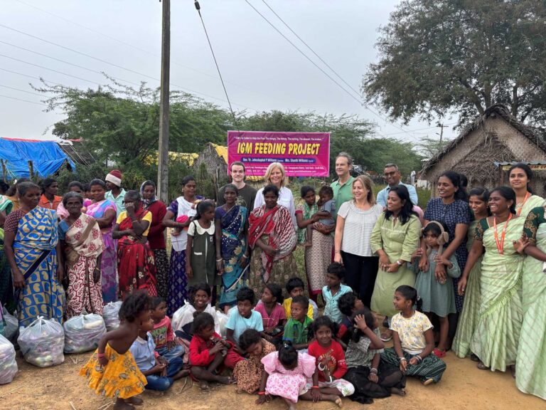 A diverse group of adults and children gather outdoors in India in front of a banner that reads IGM Feeding Project. Some people are seated, others standing, with bags of supplies nearby and simple houses and trees in the background.