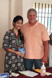 A smiling woman in a black patterned dress stands beside a man in a peach polo shirt at a table with papers, a sanitizer bottle, and a pen. They are indoors in India, with a window and bars visible in the background.