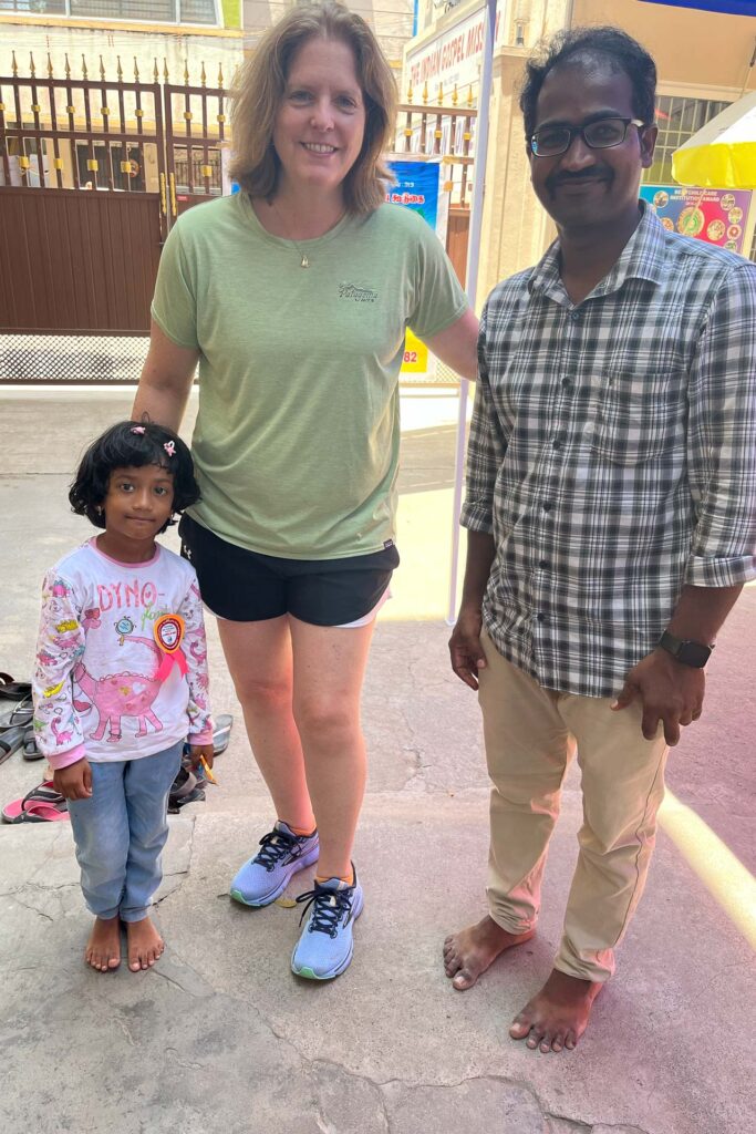 Three people—an adult woman in a green shirt and shorts, a barefoot young girl in a unicorn shirt, and a barefoot man in a plaid shirt—stand together outside on a sunny day in India, smiling at the camera.