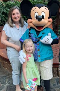 A woman and a young girl stand smiling next to a person in a Mickey Mouse costume wearing a blue Hawaiian shirt. The girl holds colorful papers. They are outdoors in India, surrounded by greenery and wooden benches in the background.