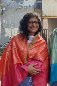 A woman with curly hair and glasses smiles while wearing a bright, patterned pink and orange shawl inspired by India, standing outdoors in front of a weathered building.