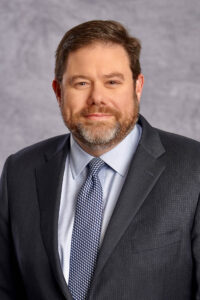 A man with short brown hair and a beard, wearing a dark suit, light blue shirt, and patterned tie, poses in front of a gray textured background, bringing a touch of India-inspired sophistication to his look.