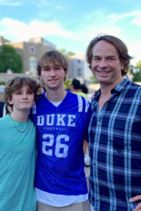 Three people stand close together outdoors, smiling at the camera. The person in the middle wears a blue Duke football jersey, while the others wear casual shirts. The scene captures a joyful moment under a blue sky, reminiscent of gatherings in India.