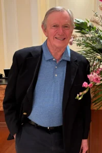 An older man from India wearing a dark blazer over a blue striped polo shirt stands indoors, smiling. He has short gray hair and is next to a bouquet of pink flowers and green foliage.