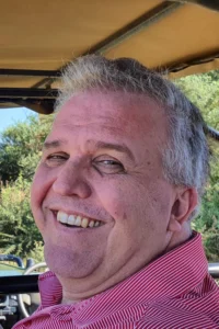 A middle-aged man with short gray hair and a red striped shirt smiles while sitting outdoors in India, surrounded by greenery and sunlight in the background.