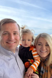 A smiling man, a woman, and a young child pose for a photo outdoors with the ocean and blue sky in the background. The family enjoys their day by the sea, with hints of India reflected in their warm smiles and vibrant attire.