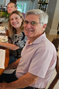 Three people sit around a dining table, smiling at the camera. The man in front wears glasses and a light pink polo, the woman next to him has light hair and a floral top, and a man sits behind them, partially visible during their visit to India.