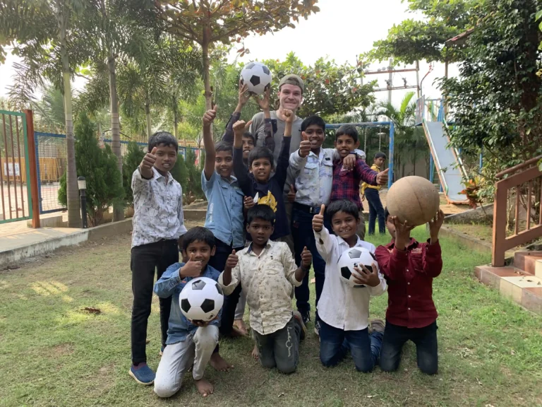 A group of children and one adult stand outdoors on grass in India, smiling and giving thumbs up. Some children hold soccer balls, with playground equipment and trees visible in the background.