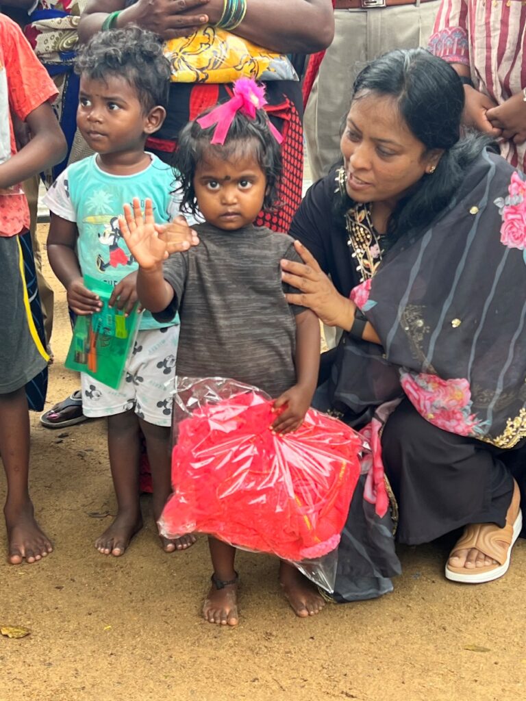 A young girl in India holding a red package stands barefoot, raising her hand, as a woman beside her gently supports her. Another small child and several people stand in the background, all outdoors on sandy ground.
