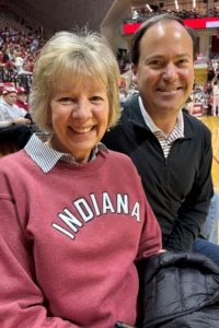 A smiling woman in an Indiana sweatshirt and a man in a dark jacket sit together at an indoor sports arena, with a crowd and basketball court in the background, sharing their love for basketball far from India.