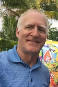 Smiling older man in a blue polo shirt stands outdoors in front of colorful artwork and palm trees, evoking the vibrant spirit of India.