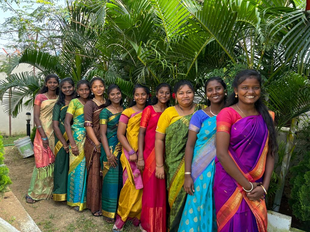 Ten women stand outdoors in a garden in India, smiling and wearing colorful saris in shades of green, maroon, yellow, pink, blue, and purple, with lush green plants and trees in the background.