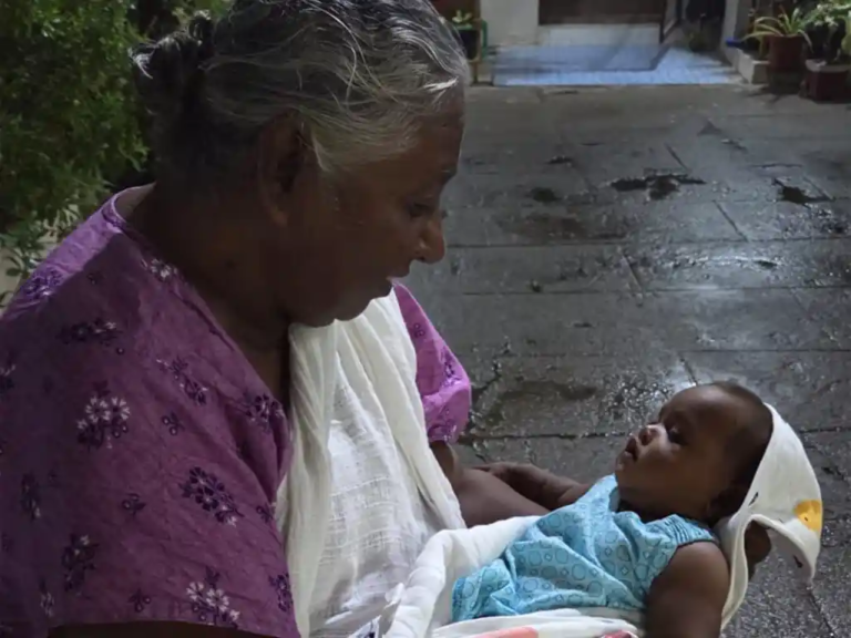 An elderly woman with gray hair holds a baby wrapped in a blanket on her lap. Sitting outside on a paved area near some plants in India, she gazes lovingly at the baby.