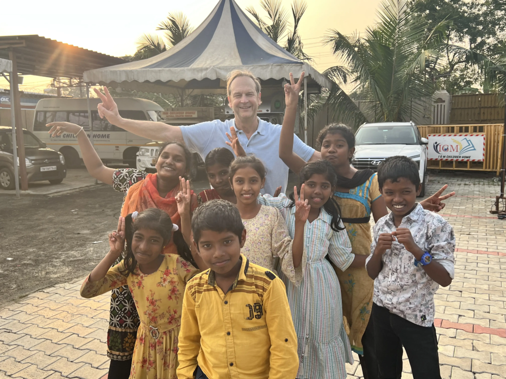 A group of smiling children and a man stand outdoors in India, posing together and making peace signs. They are in front of tents, vans, and palm trees, enjoying a sunny day.