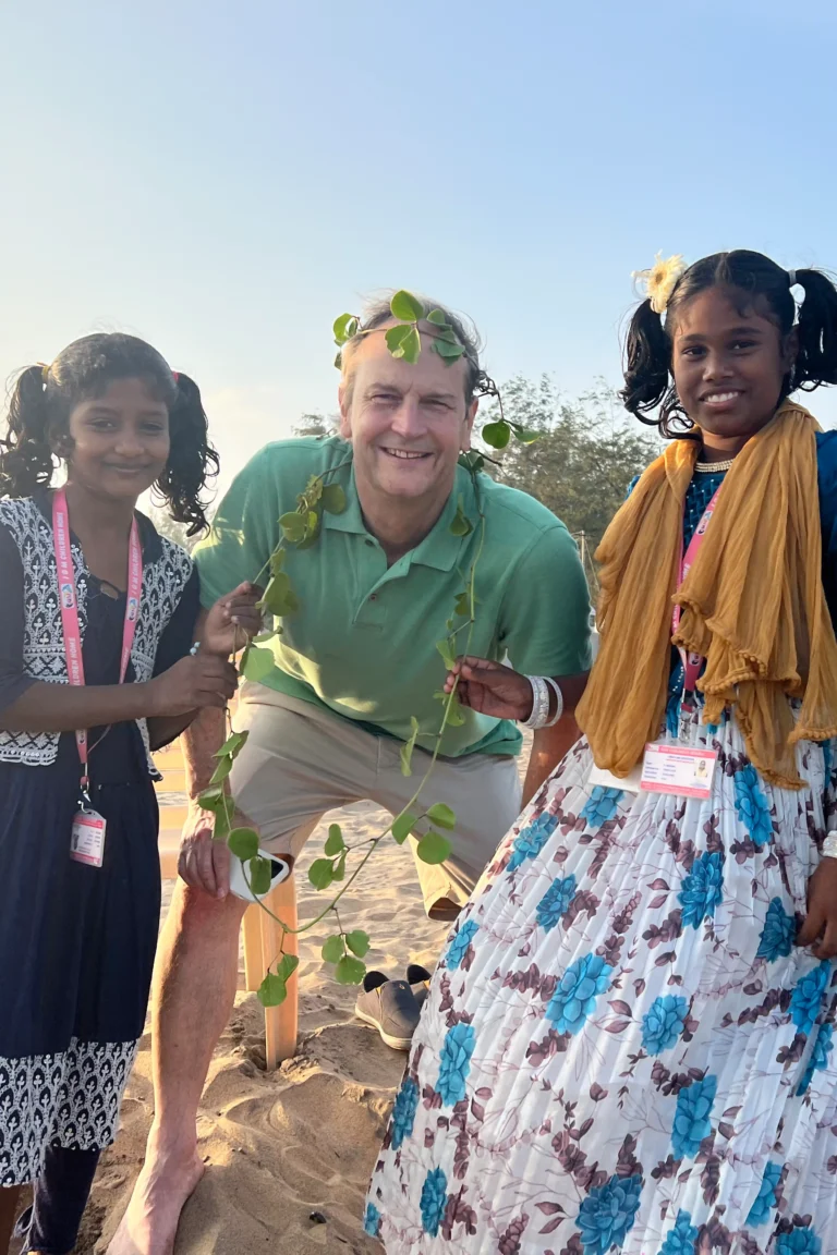 A man in a green shirt kneels on sand between two smiling girls, each holding a leafy vine. The girls, wearing colorful outfits and ID lanyards, enjoy the outdoors in India with blue sky and trees behind them.