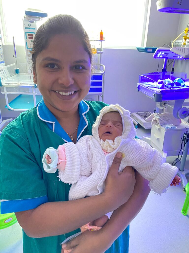 A smiling nurse in green scrubs holds a newborn baby dressed in a white knitted outfit in a hospital neonatal unit in India. Medical equipment and a lit incubator can be seen in the background.