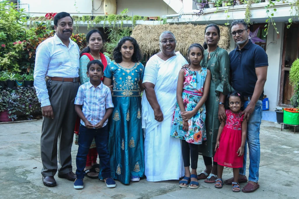 A group of nine people, including adults and children, stand together outdoors in India, smiling for a photo. Some women and girls wear colorful dresses, while others wear traditional attire. Plants and a building are visible in the background.