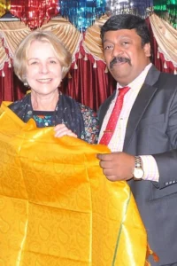 Two people smiling and holding a bright yellow patterned fabric from India, standing in front of colorful decorations and red curtains.