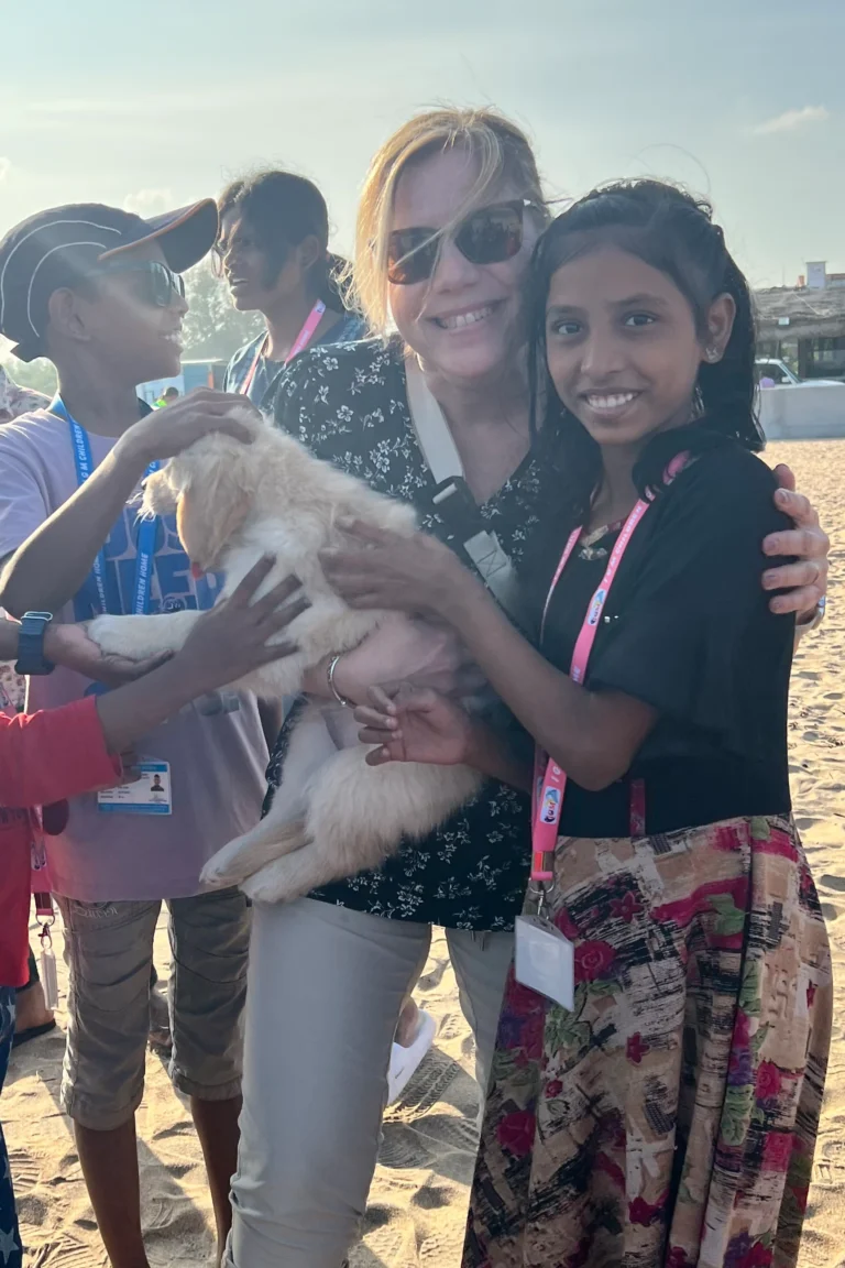A smiling woman in sunglasses stands on a sunny beach in India with children, all gathered around a fluffy puppy. The group looks happy, with buildings visible in the background.