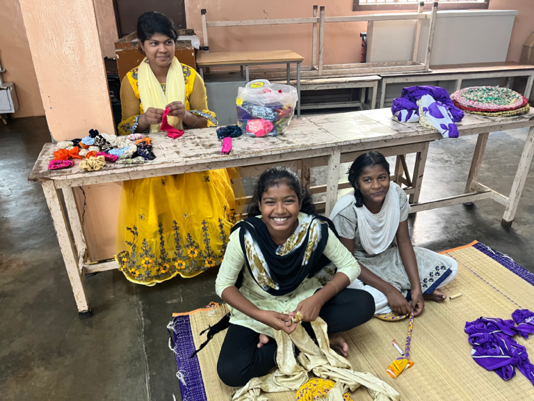 Three girls in an Indian classroom sit on the floor and at a desk, smiling while making colorful crafts. The room features peach-colored walls, with materials spread out on the desk and woven mats on the floor.