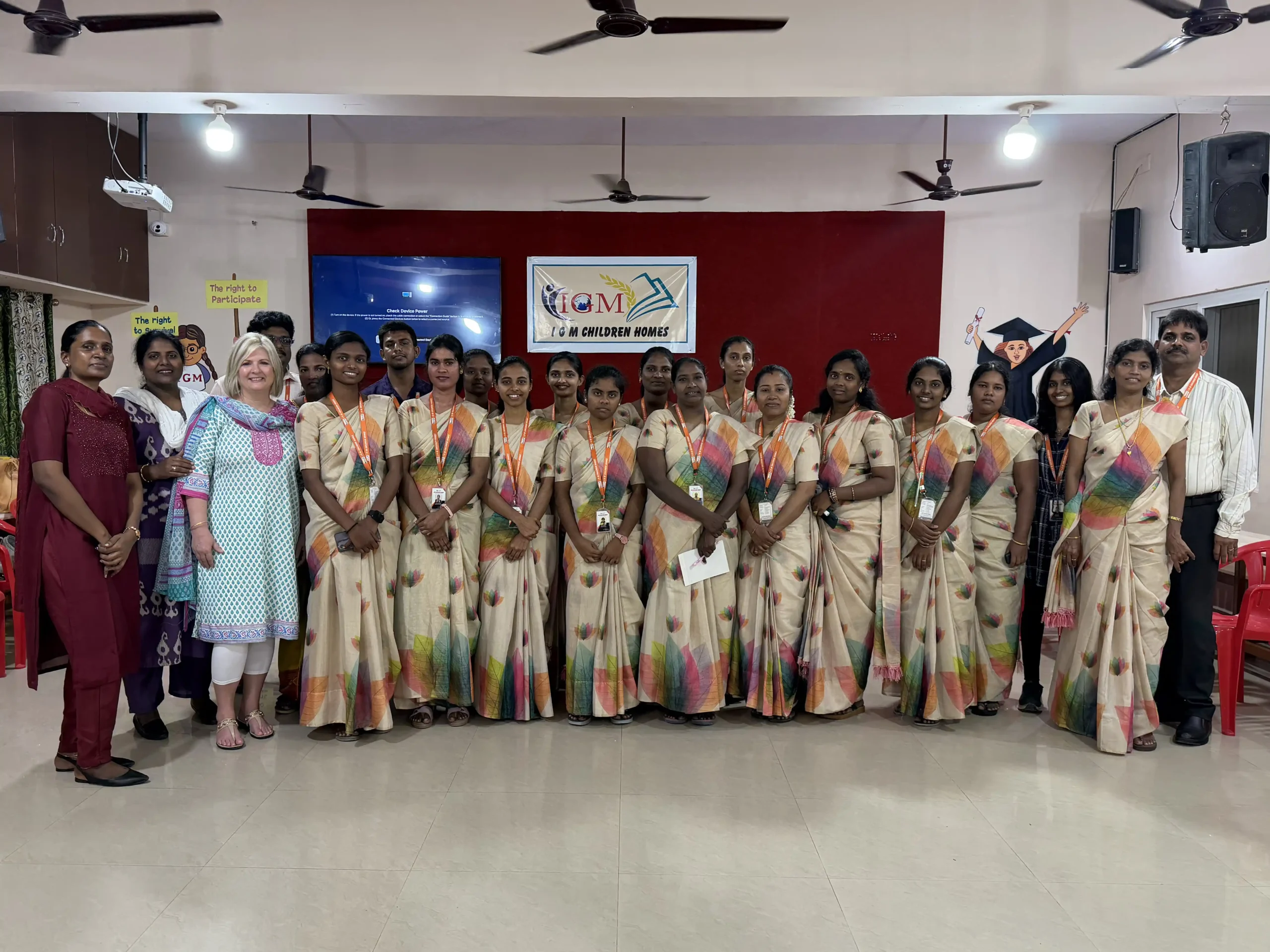 A group of women in matching cream sarees with colorful borders and a few others stand together indoors, smiling for a group photo in front of a red wall with a sign reading “ICM Children House” in India.