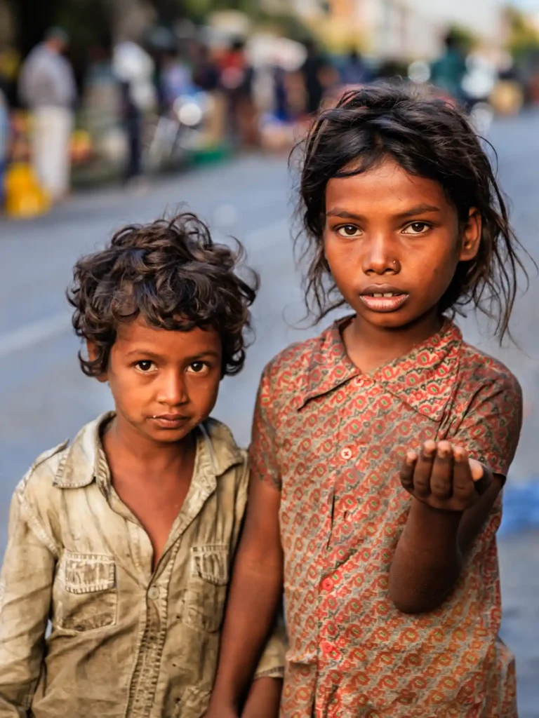 Two young children with disheveled hair and worn clothes stand on a street in India, one holding out a hand as if asking for help, with a blurred urban background behind them.