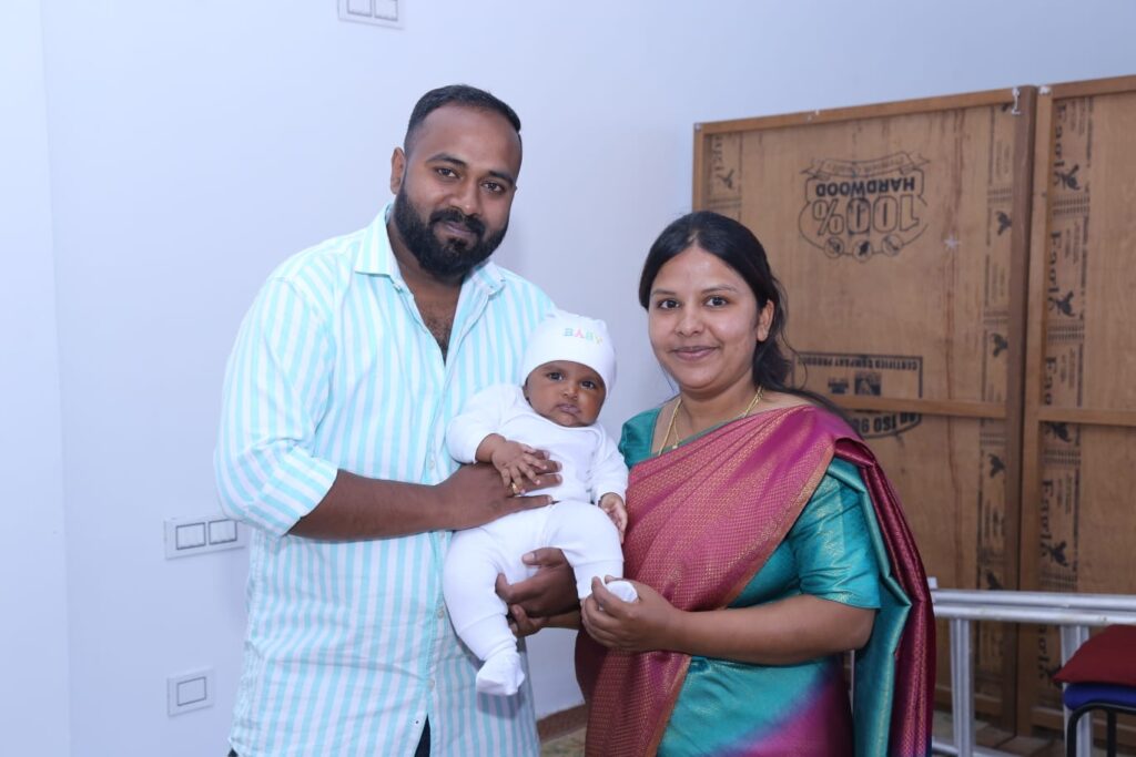 A man and woman smile at the camera while holding a baby dressed in white. Standing indoors beside a wooden panel and chairs, the woman wears a colorful saree, reflecting the vibrant traditions of India.