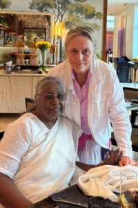 Two women pose together at an India restaurant. One is seated, wearing a white saree and glasses, while the other stands beside her in a white jacket over a pink shirt. They both smile warmly at the camera.